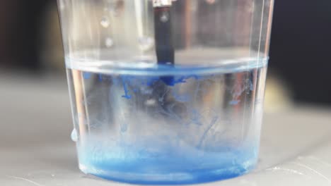 Ein-Tropfen-Blauer-Farbe-Tropft-Und-Diffundiert-Zu-Ranken-In-Einem-Glas-Wasser-Und-Reinigt-Den-Aquarellpinsel