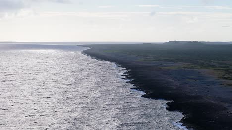 Aerial-trucking-pan-across-black-volcanic-rock-spreading-into-open-ocean-water