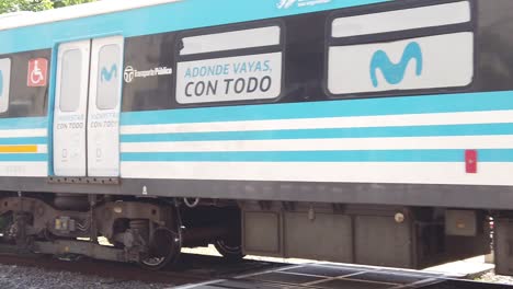 Public-Transport-Blue-Train-in-Slow-Motion-Drives-Through-Sarmiento-Railway-City-with-Movistar-Ads,-Flores-Neighborhood