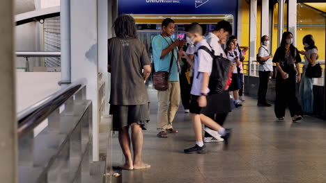 Un-Hombre-Sin-Hogar-Está-Pidiendo-Limosna-Mientras-Los-Peatones-Pasan-Por-Un-Puente-Peatonal-Frente-A-La-Entrada-Del-Union-Mall-En-Bangkok,-Tailandia