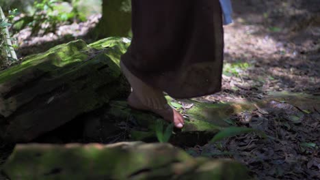 Brunette-woman-walking-on-the-slatted-stones-of-a-lush-forest,-with-bare-feet-wearing-a-brown-dress
