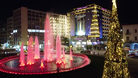 Árbol-De-Navidad-Con-Fuente-De-Agua-En-La-Plaza-Omonoia-Y-El-Edificio-Del-Centro-Hondos-Al-Fondo-Por-La-Noche-En-Atenas,-Grecia
