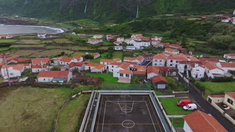 Fly-above-soccer-court-at-Fajã-Grande-town-with-waterfall-in-background