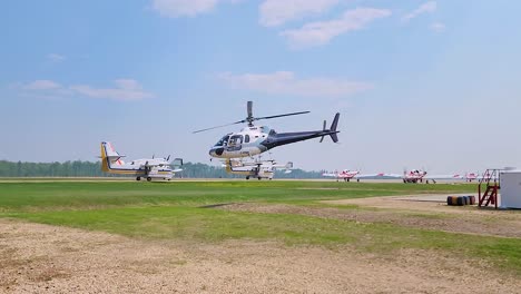 A-white-helicopter-is-taking-off-from-a-grassy-field-with-small-planes-and-equipment-in-the-background-under-a-clear-sky