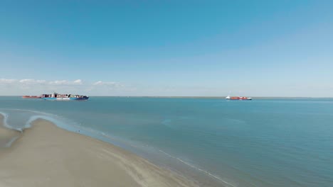 An-aerial-view-captures-a-loaded-container-ship-passing-by-the-east-end-of-Galveston-Island-while-a-couple-walks-down-the-beach-under-clear-blue-skies-in-Galveston,-Texas