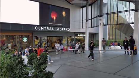Käufer-Gehen-Im-Zentralen-Einkaufszentrum-Ladprao-Ein-Und-Aus,-Während-Einige-Umstehende-Auf-Den-Bänken-Am-Eingang-Des-Kaufhauses-In-Bangkok,-Thailand,-Sitzen