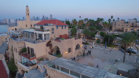 Push-in-shot-over-the-beautiful-and-old-buildings-of-the-Jaffa-Old-city-at-c-with-people-sitting-on-the-roofs-to-watch-the-sunset---Tel-Aviv-towers-and-the-old-Kedumim-square-in-the-background