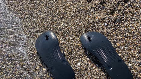Waves-hitting-washed-up-black-upside-down-flip-flops-on-a-beach-with-tiny-little-rocks-in-Spain,-sunny-summer-vacation-holiday-shoes,-4K-shot