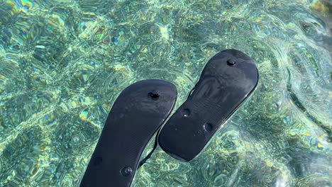 Floating-black-upside-down-flip-flops-in-transparent-turquoise-sea-water-by-the-beach-in-Spain,-fun-sunny-summer-vacation-holiday-shoes,-4K-shot