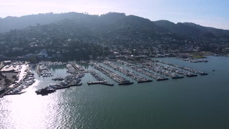 Drone-view-over-Sausalitoâ€™s-bustling-marina,-yachts-moored-in-neat-rows,-bordered-by-verdant-hills,-with-a-hint-of-San-Francisco-afar