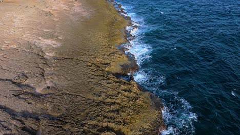Drone-bird's-eye-view-dolly-along-green-coastline-bombarded-by-strong-ocean-waves-eroding-away-basalt
