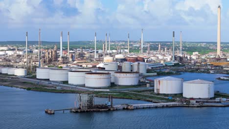 Oil-refinery-stacks-and-storage-tanks-on-coastal-tropical-island-covered-by-cloud-shadow