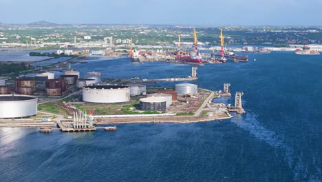 Aerial-parallax-around-oil-refinery-stack-and-storage-vats-on-coast-of-tropical-island