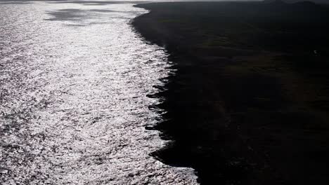 Harsh-light-spread-across-ocean-shimmering-against-dark-basalt-rock-on-island-shoreline