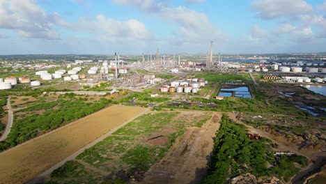 Aerial-trucking-pan-above-empty-grass-green-space-with-oil-refinery-flare-stacks-in-distance