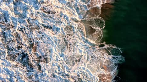 Aerial-view-of-the-foamy-waves-crashing-against-the-shoreline,-Sunlight-casting-a-golden-glow-on-the-ocean's-froth