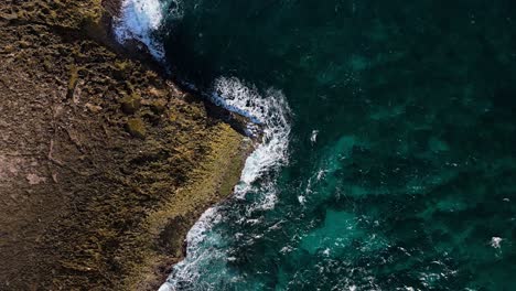 Drone-top-down-descends-on-jagged-sharp-rocky-ocean-point-with-view-of-reef-below-clear-water