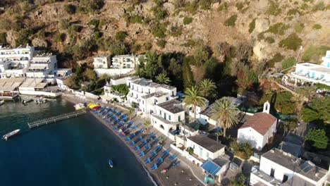 Drone-view-in-Greece-flying-over-blue-sea-in-Loutro-small-white-house-town-and-small-boats-next-to-a-hill-on-a-sunny-day
