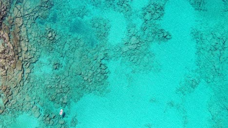 Drone-view-in-Greece-top-view-ocean-clear-blue-water-with-rocks-on-the-bottom-a-small-white-boat-on-a-sunny-day