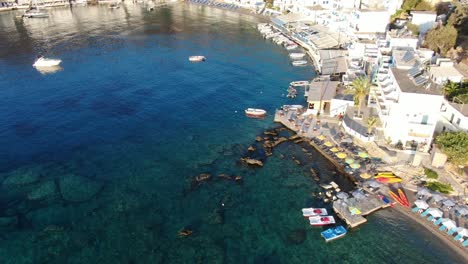 Drone-view-in-Greece-flying-over-blue-sea-in-Loutro-small-white-house-town-and-small-boats-next-to-a-hill-on-a-sunny-day
