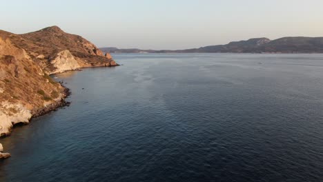 Drone-view-in-Greece-flying-over-blue-sea-next-a-brown-hill-mountain-at-sunset-in-Milos