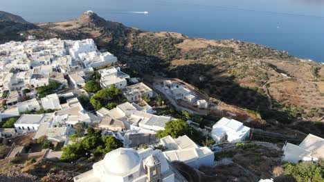 Drone-view-in-Greece-vertical-ascend-over-a-white-church-on-a-hill-with-a-greek-white-house-town-facing-blue-sea-on-a-mountain