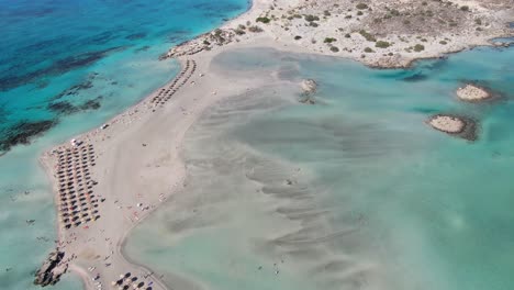 Drohnenansicht-In-Griechenland,-Die-An-Einem-Sonnigen-Tag-über-Den-Schmalen-Weißen-Sandstrand-Von-Elafonisi-Fliegt,-Klares-Blaues-Wasser-An-Den-Seiten-Und-Viele-Sonnenschirme