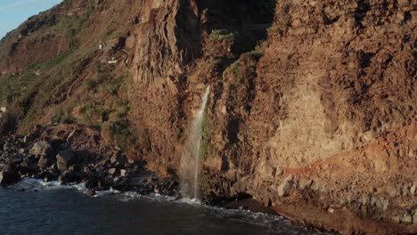 Small-beautiful-waterfall-pouring-into-ocean-from-mountain-wall-with-waves-crashing-against-during-sunset-in-Ponta-Do-Sol,-Madeira,-Portugal