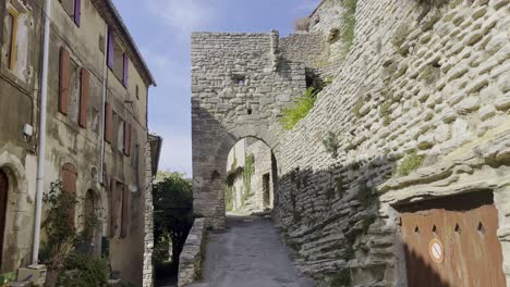Small-street-in-a-historic-village-with-old-stone-wall-rest-with-archway-to-drive-through-in-the-sunshine-of-France