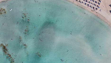 Drone-view-in-Greece-top-view-ocean-clear-blue-water-with-rocks-on-the-bottom-and-a-narrow-white-sand-beach-with-umbrellas-on-a-sunny-day