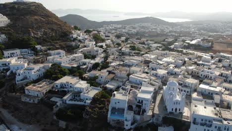 Drohnenansicht-In-Griechenland,-Die-An-Einem-Sonnigen-Tag-Vor-Einer-Griechischen-Stadt-Mit-Weißen-Häusern-Auf-Einem-Braunen-Hügel-Und-Dem-Meer-Am-Horizont-Fliegt