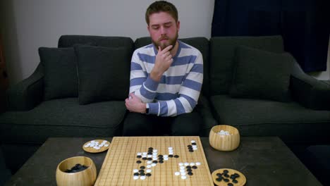 Beaded-man-strokes-beard-while-thinking-of-strategic-move-in-Go-board-game-on-bamboo-board-on-stone-table-by-grey-couch-in-living-room