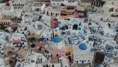 Drone-view-in-Greece-flying-over-Santorini-with-Oia-town-white-houses-at-close-with-colored-fronts-and-pools-at-sunrise