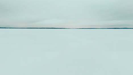 Flying-over-a-road-on-frozen-lake-ice-to-reveal-a-snowy-forest-in-the-horizon