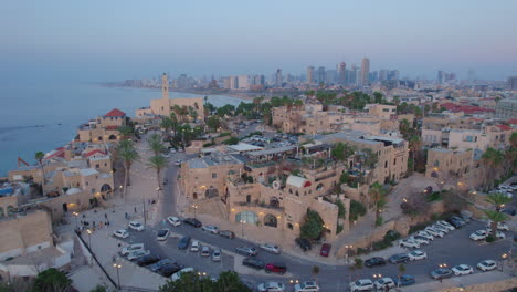 Cloes-parallax-drone-shot-of-Old-city-of-Jaffa-and-Jaffa-port-at-sunset-with-lots-of-families-visiting-restaurants,-shops-and-bars-in-the-port---tel-aviv-city-at-the-background
