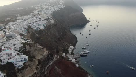 Drone-view-in-Greece-flying-over-Santorini-with-Oia-town-white-houses-on-a-cliff-next-to-the-mediterranean-sea-at-sunrise