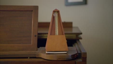 An-antique-vintage-wooden-metronome-set-to-42-beats-per-minute-swings-slowly-back-and-forth-to-mark-time-on-top-of-an-old-brown-piano