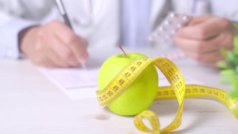 Nutrition-appointment-concept,-Doctor-holding-medicine-blister,-an-apple-and-measure-tape-in-foreground