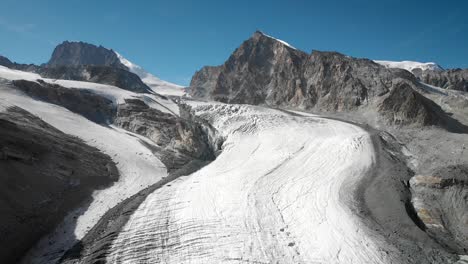 Aerial-flyover-over-the-Allalin-glacier-near-Saas-Fee-in-Valais,-Switzerland-with-a-pan-down-view-from-Allalinhorn-peak-down-to-the-crevasses-in-the-ice-on-a-sunny-summer-day-in-the-Swiss-Alps