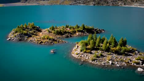 Aerial-flyover-over-forested-islands-with-sunlit-trees-in-the-turquoise-waters-of-Lac-de-Salanfe-in-Valais,-Switzerland-on-a-sunny-autumn-day-in-the-Swiss-Alps