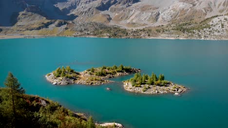 Aerial-view-of-tiny-islands-in-the-waters-of-Lac-de-Salanfe-in-Valais,-Switzerland-on-a-sunny-autumn-day-in-the-Swiss-Alps-with-a-pan-up-view-to-surrounding-alpine-peaks-and-cliffs