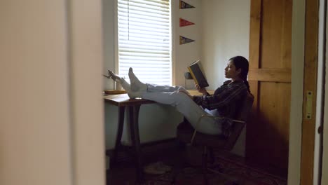 Relaxed-Puerto-Rican-female-reading-captivating-story-book-with-feet-raised-on-bedroom-desk