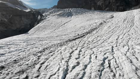 Sobrevuelo-Aéreo-Sobre-Un-Excursionista-Caminando-Sobre-El-Hielo-Del-Glaciar-Allalin-Cerca-De-Saas-fee-En-Valais,-Suiza,-Mientras-El-Sol-Brilla-Detrás-De-Los-Picos-De-Los-Alpes-Suizos