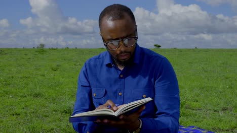 A-black-man-sits-in-the-grass-and-intently-studies-a-book-with-pen-in-hand