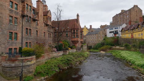 Slow-motion-pan-across-flowing-river-bubbling-on-mossy-shores-between-tall-old-brown-brick-buildings-of-Edinburgh-Scotland