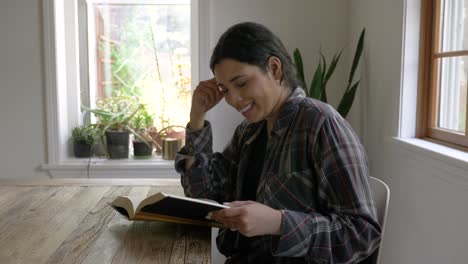 Mujer-Multiétnica-Sonriente-Sentada-Leyendo-Un-Libro-Relajado-En-El-Interior-De-Una-Casa