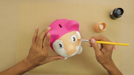 Painting-plaster-piggy-bank-with-paintbrush-on-brown-paper-at-home