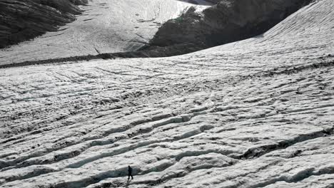 Aerial-flyover-over-a-hiker-walking-across-the-ice-of-the-Allalin-glacier-near-Saas-Fee-in-Valais,-Switzerland-on-a-sunny-day-in-the-Swiss-Alps-with-a-pan-up-view-from-the-crevasses-up-to-the-peaks