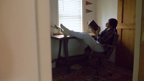 Inquisitive-Puerto-Rican-female-reading-captivating-book-with-feet-raised-on-home-office-desk