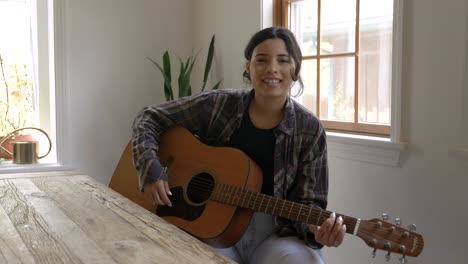 Happy-young-adult-Puerto-Rican-female-playing-acoustic-guitar-and-singing-in-home-interior